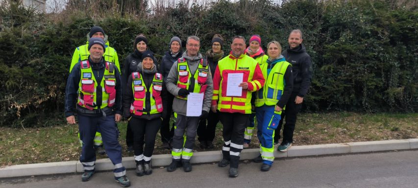Gruppe von Rettungskräften in Uniform steht lächelnd vor einer Hecke.